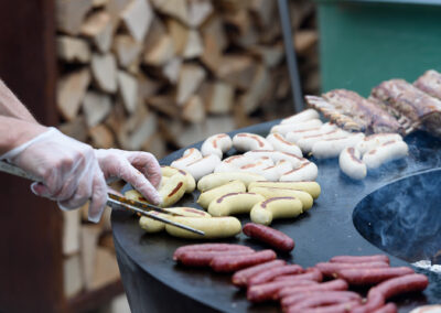 Eine Person mit Handschuhen schneidet verschiedene Wurstsorten auf einem Grill. Im Hintergrund sind Holzscheite sichtbar.